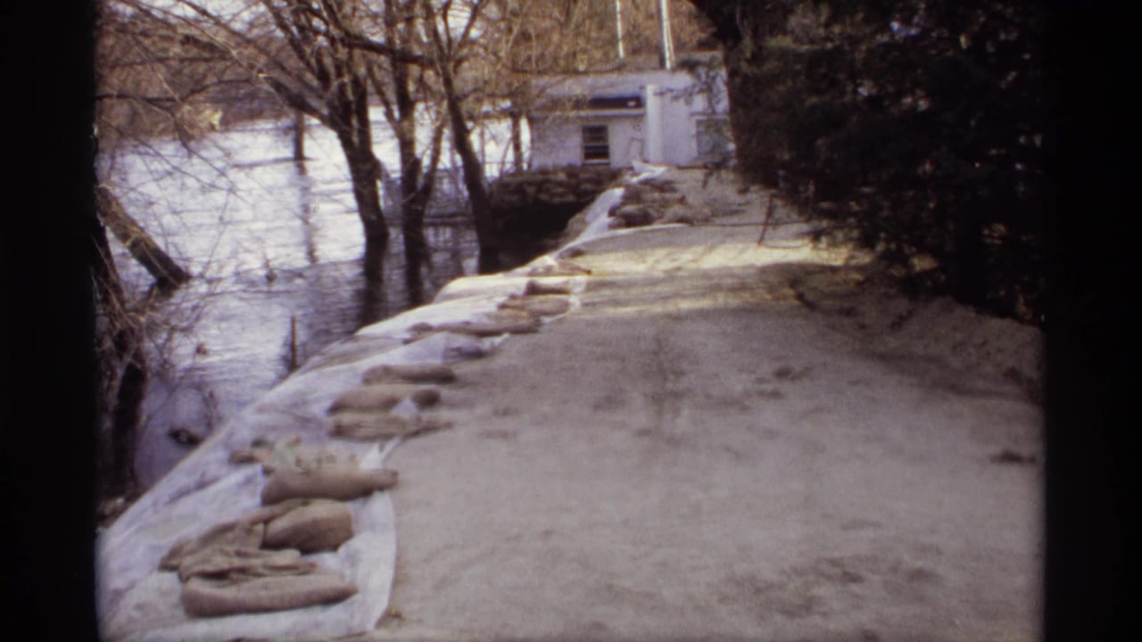 1969 Rockford, Minnesota 8mm frame showing floodwater along a sandbagged road with bare trees and a small building beyond.
