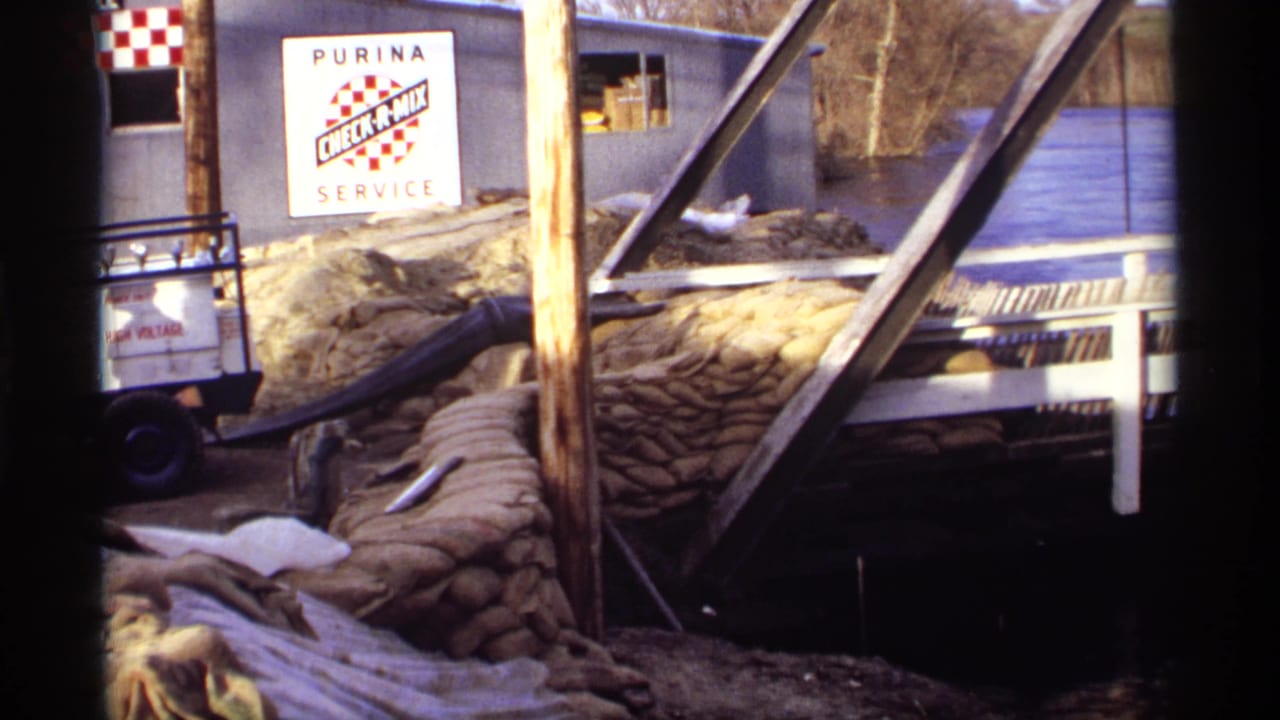 1969 Rockford, Minnesota 8mm frame showing a sandbagged riverside building, Purina Check-A-Mix Service sign, and floodwater near a bridge.