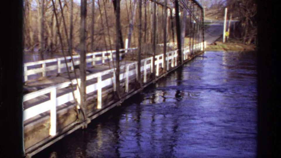 1969 Rockford, Minnesota home-movie frame showing floodwater below a wooden bridge, with white railings and bare trees on the riverbank.