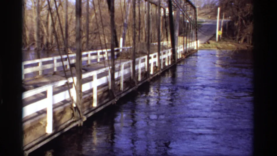 1969 Rockford, Minnesota 8mm frame showing a wooden truss bridge, white railings, and high river water flowing toward a road.