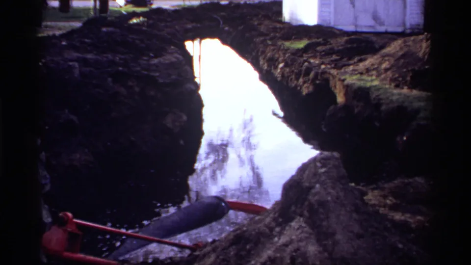1969 Rockford, Minnesota 8mm frame showing a narrow water-filled trench bordered by dark soil with a red-handled tool nearby.