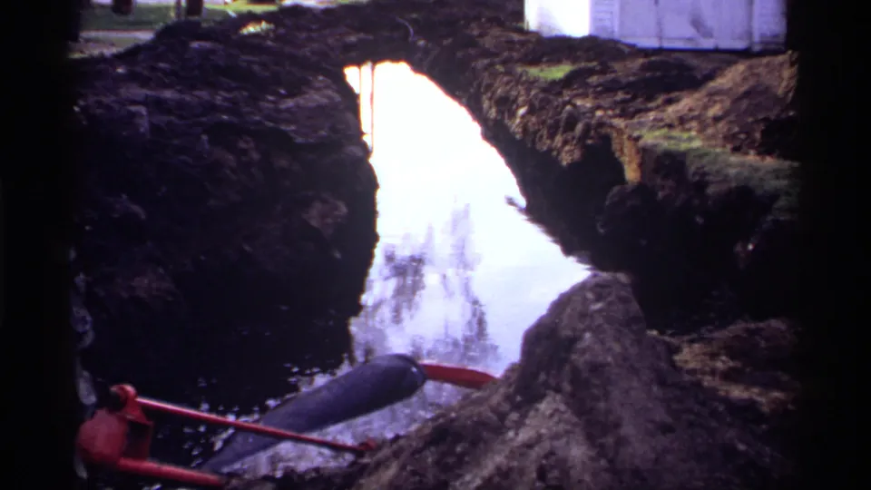 1969 Rockford, Minnesota 8mm frame showing an excavated, water-filled trench with muddy banks and a red-handled tool in the foreground.