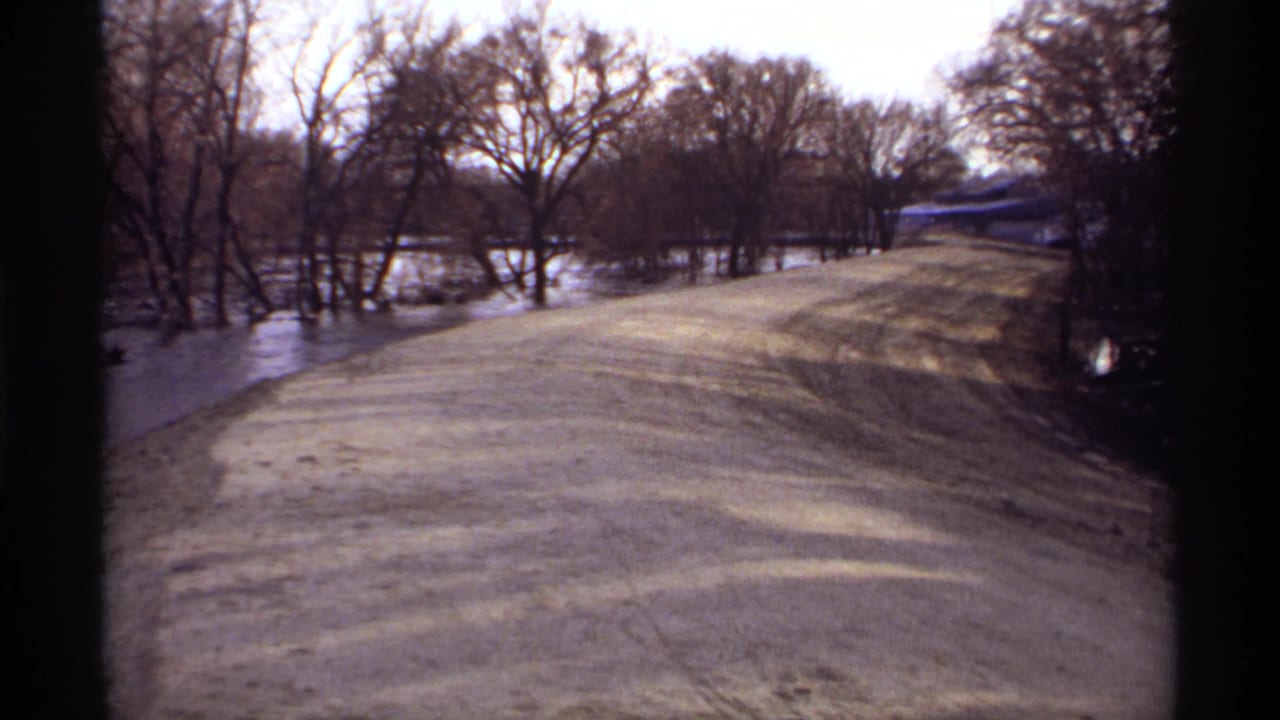 1969 8mm view of a flooded rural road in Rockford, Minnesota, with water along both sides and leafless trees in the background.