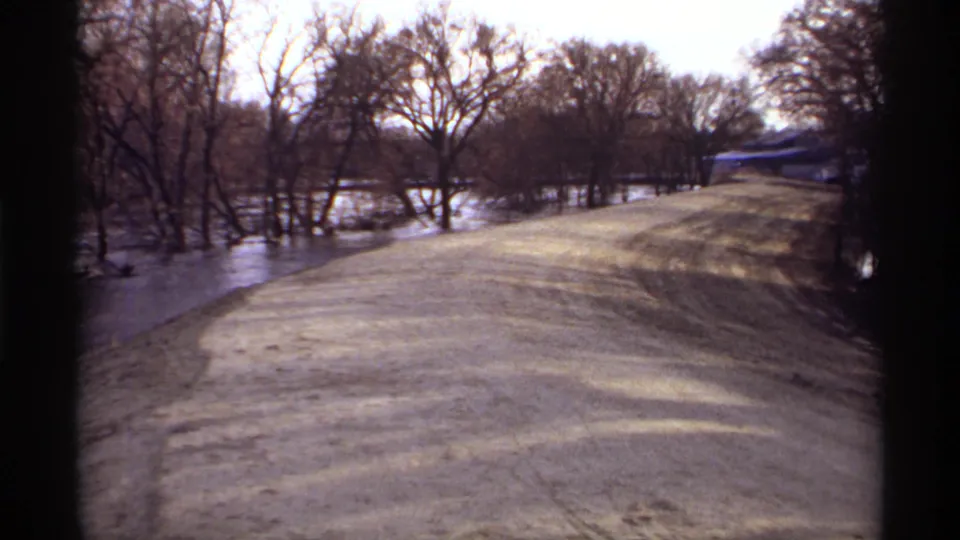Rockford, Minnesota flood scene on 8mm film, showing a dirt road with standing water on both sides and winter-bare trees beyond.