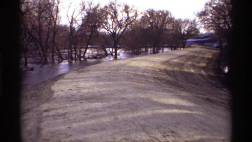 Home-movie frame from 1969 Rockford, Minnesota, showing floodwater beside a raised unpaved road under bare trees.