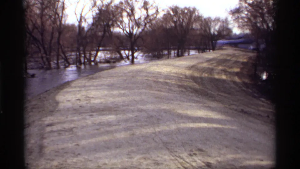 Late-1960s 8mm frame of a flooded rural road in Rockford, Minnesota, with water close to the road edge and leafless trees behind it.