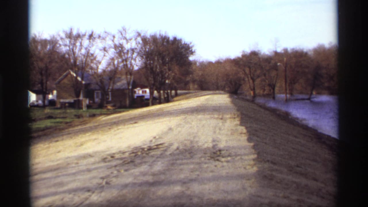 Dirt road beside a lake with leafless trees and small homes under a pale sky.