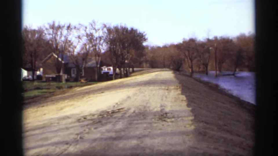 1969 Rockford, Minnesota 8mm frame showing a dirt road along calm water, with leafless trees, modest homes, and utility poles.
