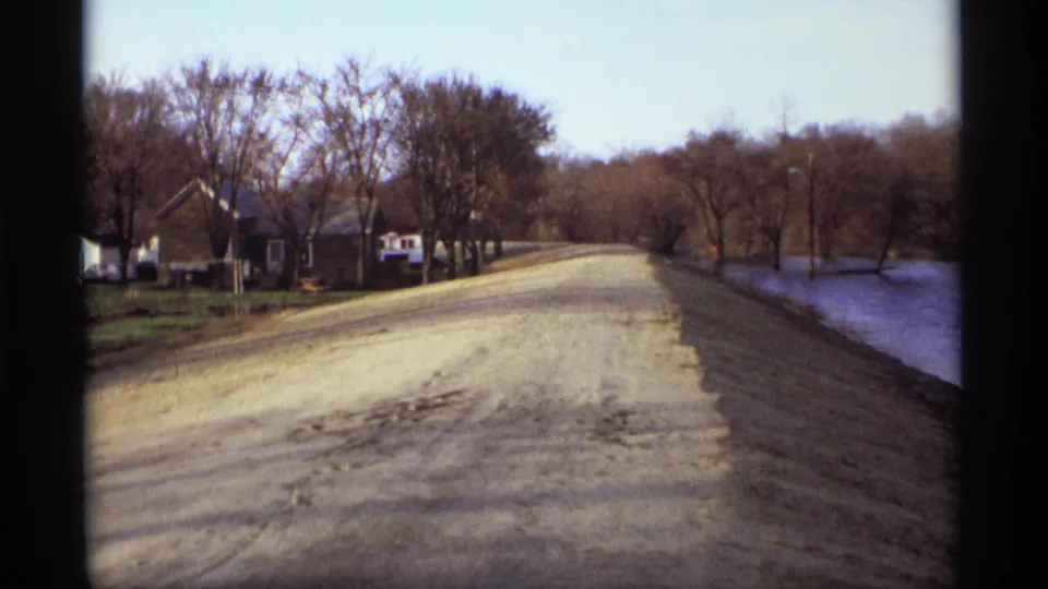 1969 Rockford, Minnesota home-movie frame with a pale dirt road, lakeshore, bare trees, and modest homes at the roadside.
