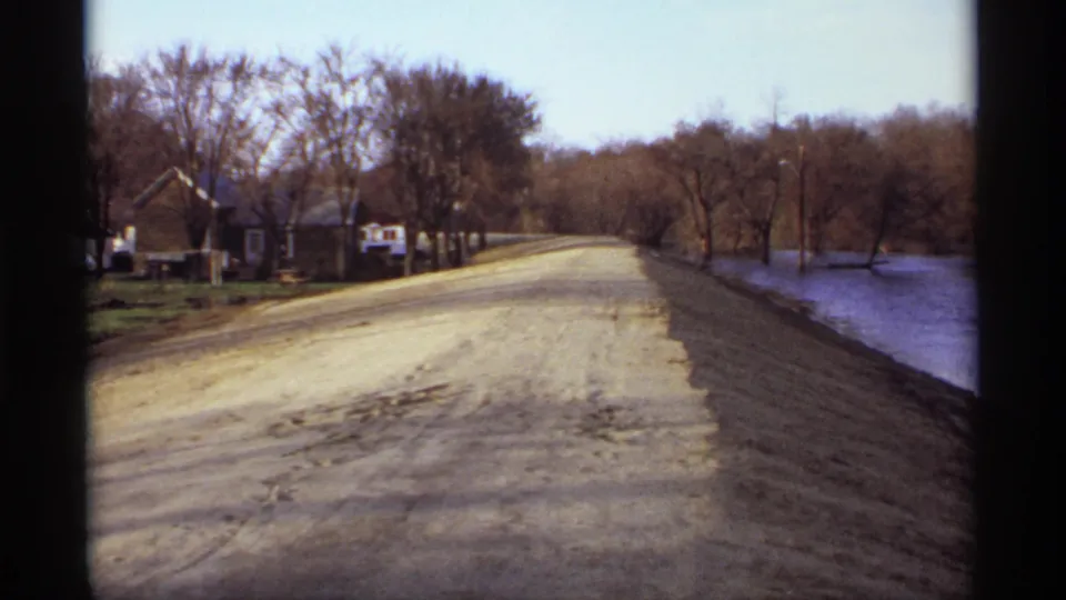 1969 Rockford, Minnesota frame showing a lakeside dirt road bordered by calm water, leafless trees, utility poles, and home exteriors.