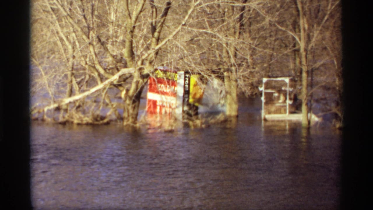 Floodwater surrounds bare trees, a red sign, and a small white structure near a residential area.