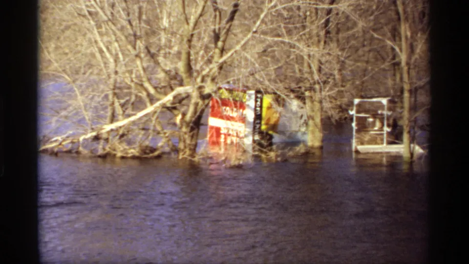 1969 Rockford, Minnesota 8mm still of high water around leafless trees, a partly submerged red sign, and a white structure.