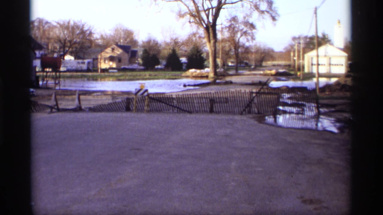 Flooded residential street with wooden barrier, houses, bare trees, and utility poles.