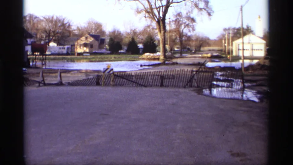 1969 Rockford, Minnesota street view with standing water, a leaning wooden barrier, houses, bare trees, and winter-bare roadside detail.