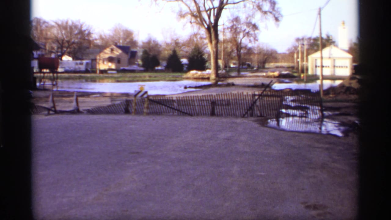 Residential street with floodwater, fence barrier, bare trees, houses, and roadside poles.