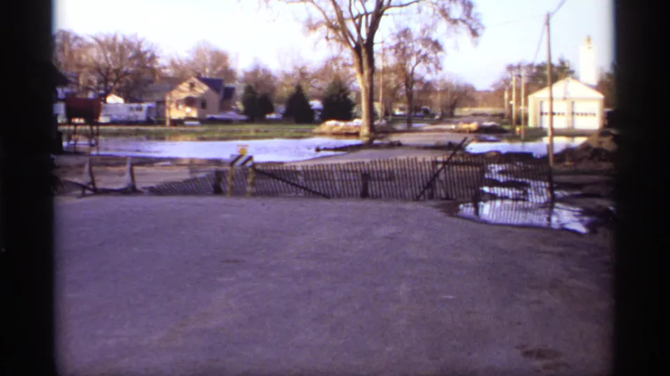 Rockford, Minnesota flood scene with a waterlogged street, wooden barrier, detached houses, leafless trees, and utility poles in soft 8mm color.