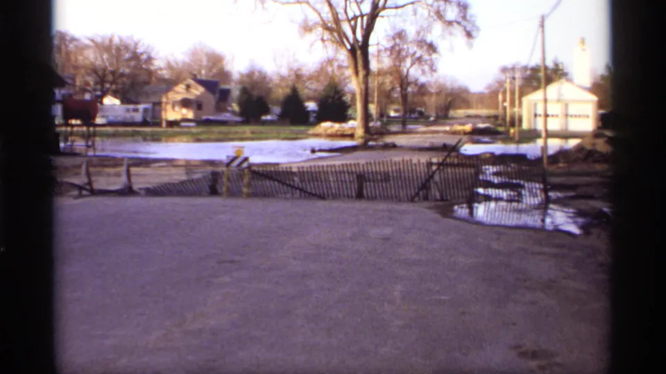1969 home-movie view of a Rockford street partly covered by floodwater, with a sagging wooden barrier, houses, trees, and roadside puddles.