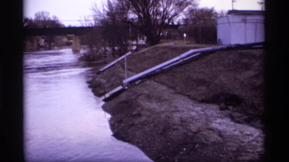 Wide 1969 Rockford, Minnesota home-movie view of a riverbank, pipes along the slope, bare trees, and a bridge beyond the water.