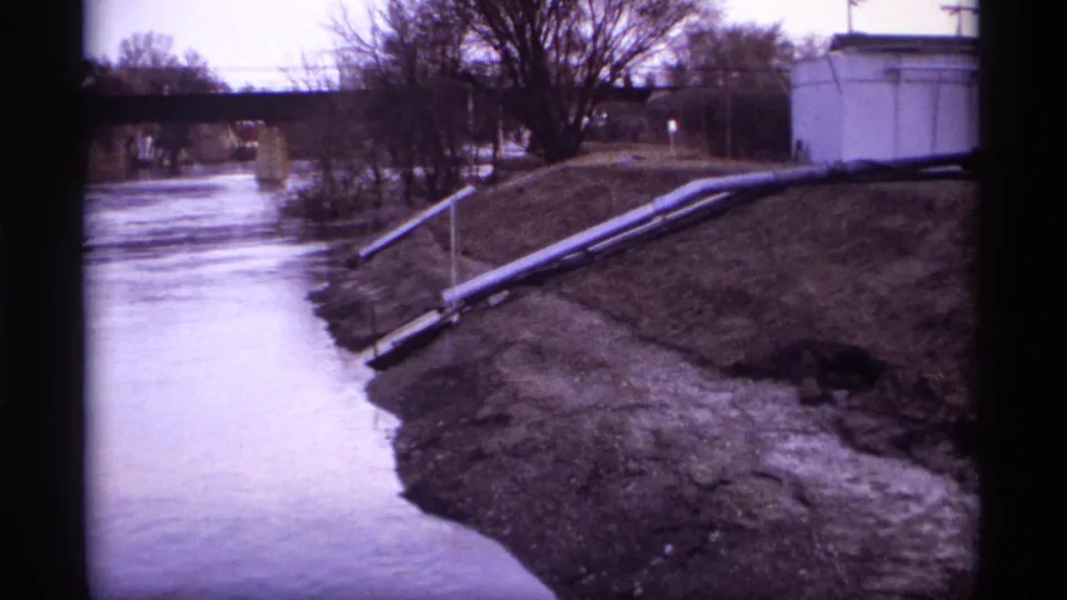 Final 8mm frame holds on the Rockford riverbank, showing muddy shoreline, exposed pipes, leafless trees, and a bridge in the background.