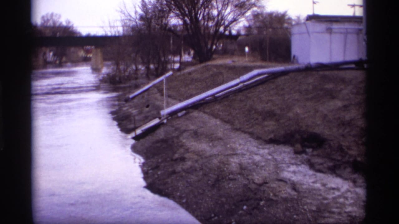 Flowing river and sloped bank with pipes, bare trees, and utility structures in the background.