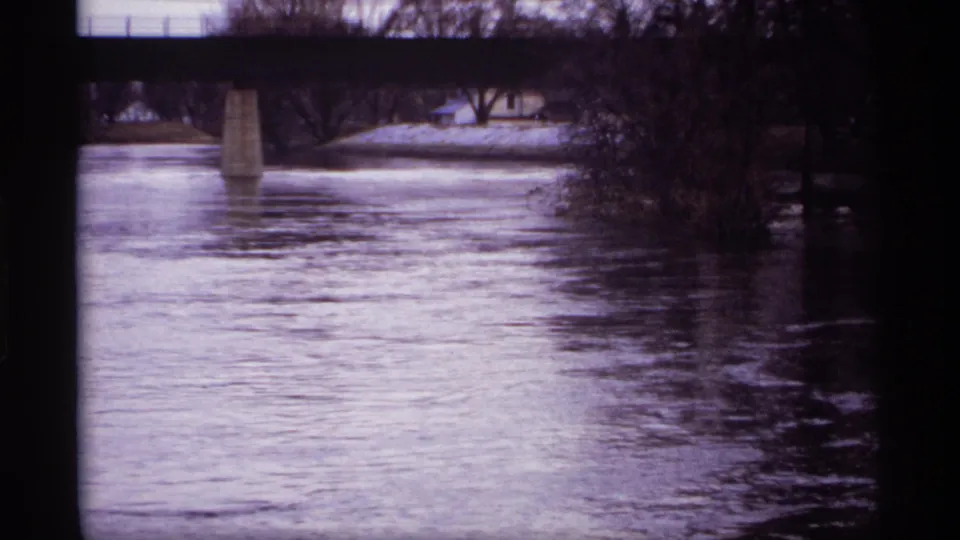 1969 8mm view of a Rockford, Minnesota river passing beneath a bridge, with a concrete pier and leafless trees along the banks.