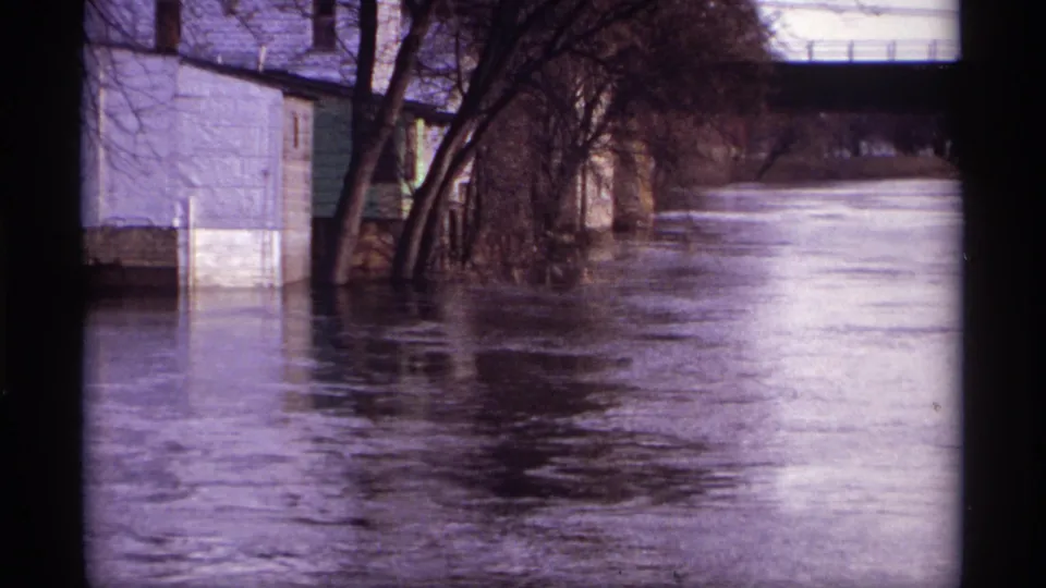 A 1969 Rockford, Minnesota home-movie frame shows high water against a riverside house, trees, and a bridge overhead.