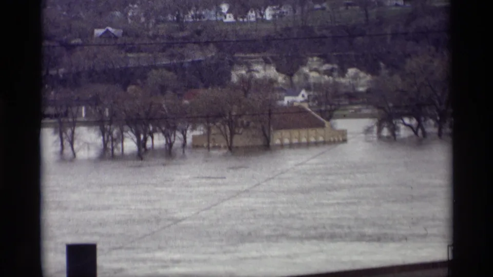 Saint Paul flood scene on 8mm film, with murky water surrounding trees and a low riverside building under a muted sky.