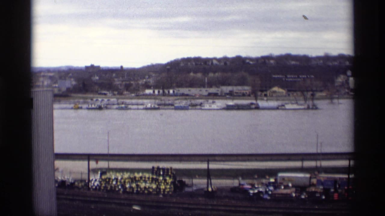 Elevated view of a riverfront with docked boats, industrial buildings, tracks, cars, and a sand pile below.