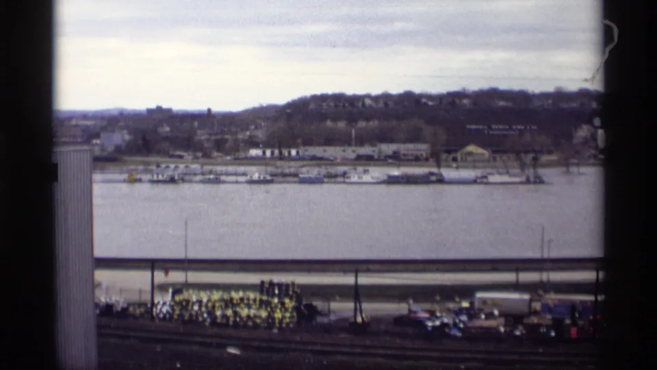 1969 Saint Paul riverfront seen from above, showing docked boats across the water, a tree-lined hill, industrial shoreline, tracks, cars, and a sand pile.