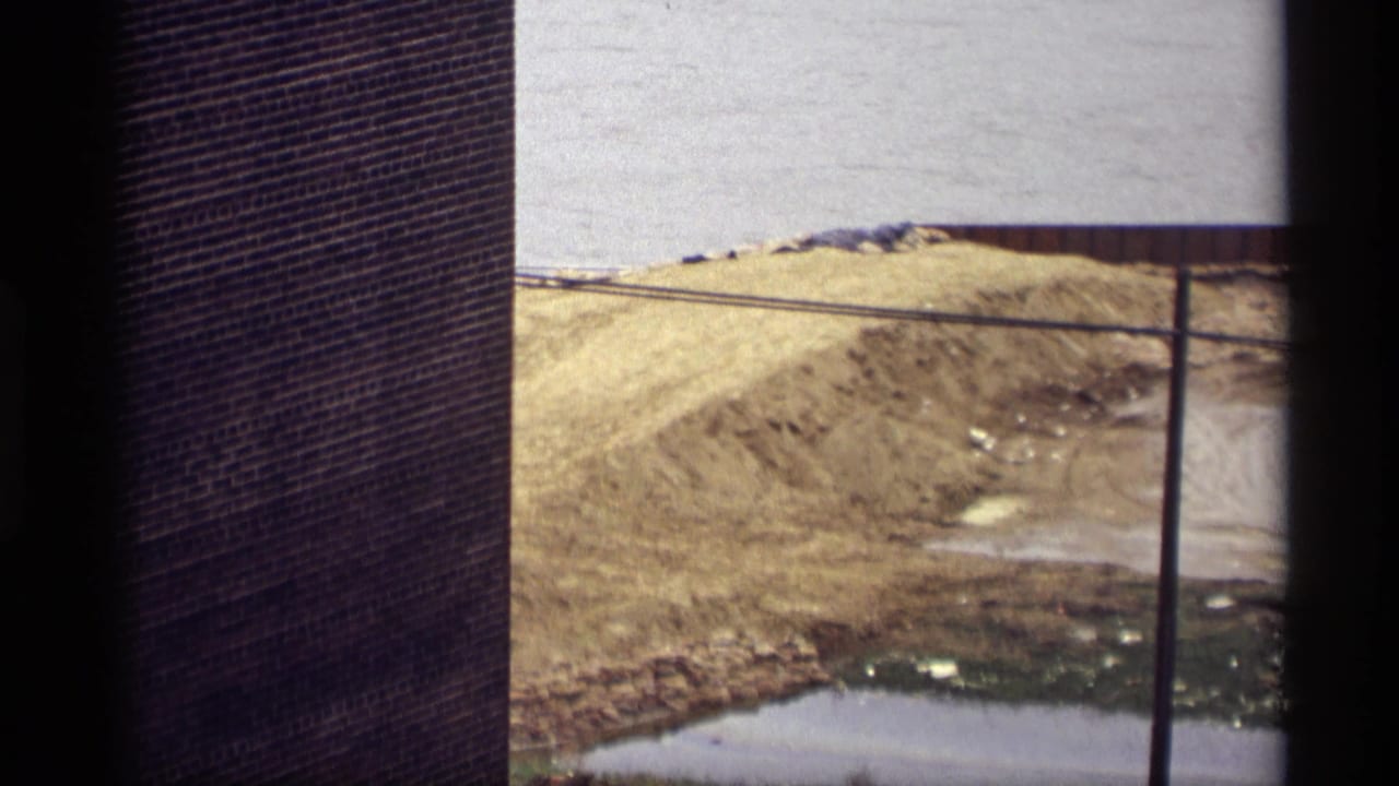 1969 Saint Paul 8mm view of a brick building edge, muddy shoreline, dirt mound, and water from an elevated position.
