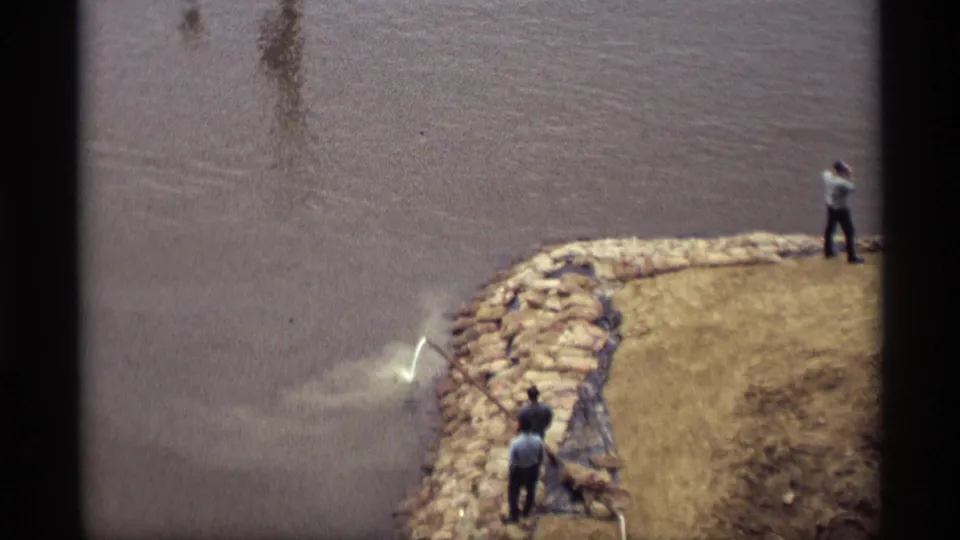 Several adult workers are seen from above on a stone-lined riverbank in Saint Paul, with hose spray visible at the waterline.