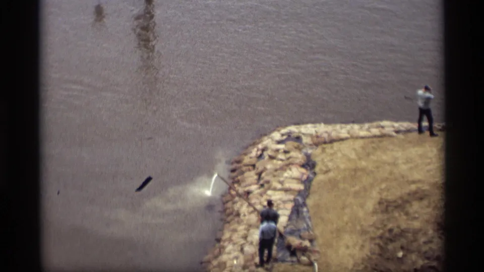 End frame shows hose spray at a muddy Saint Paul river edge, with adult workers positioned along the stone embankment and grass bank.