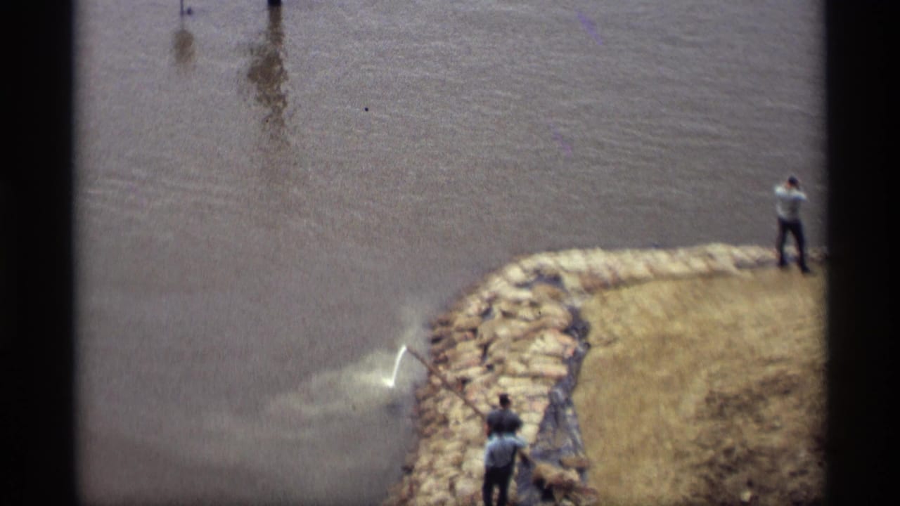 1969 Saint Paul 8mm frame showing workers beside a flooded riverbank reinforced with sandbags and dark sheeting.