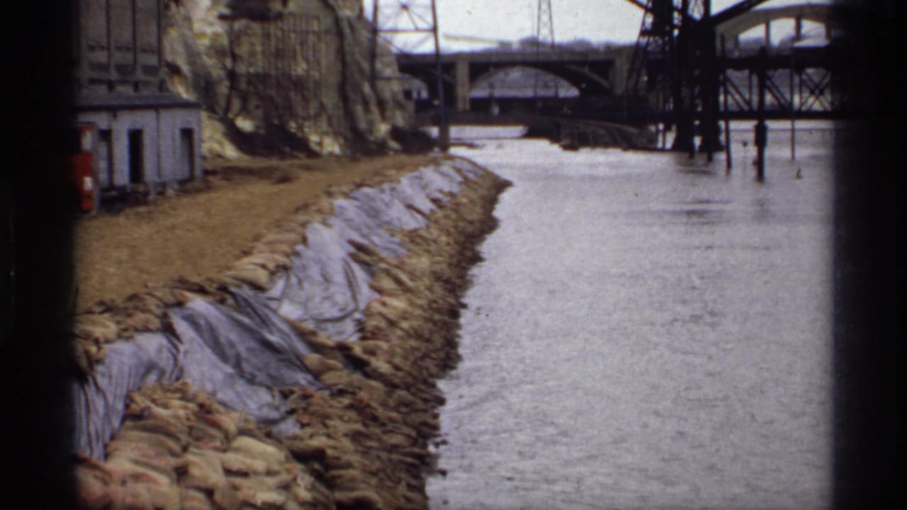 Floodwater runs beside a sandbagged shoreline with bridges in the background.