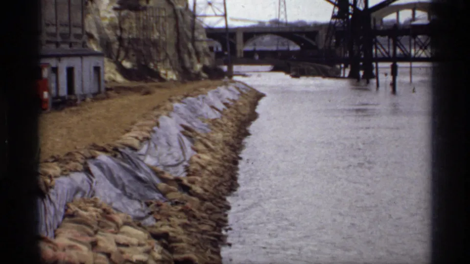 1969 Saint Paul 8mm frame of a flooded river beside a sandbagged and tarp-covered embankment near bridge structures.