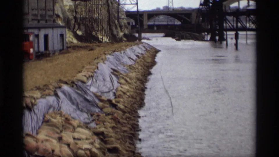 1969 Saint Paul 8mm frame showing a small riverside building near a sandbagged embankment and swollen river.