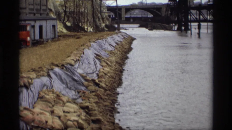 1969 Saint Paul 8mm frame of sandbags and tarp protecting a riverbank while floodwater rises beside bridge pilings.