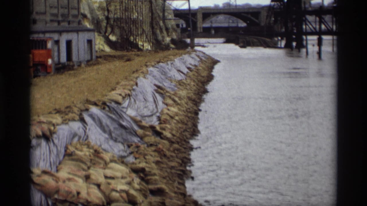 1969 Saint Paul riverfront view with sandbags, dark bank sheeting, industrial structures, and bridges spanning the water.