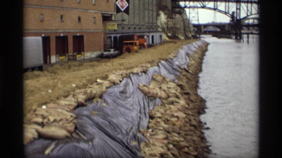 Saint Paul industrial riverbank with a brick warehouse, orange truck, sandbags, dark sheeting, and bridge structures in view.