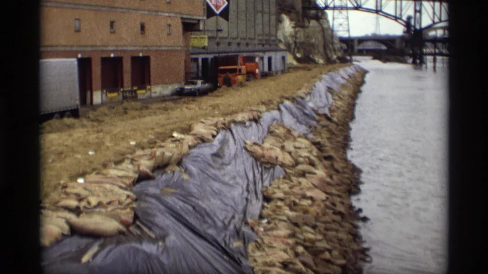 Close 1969 Saint Paul waterfront view of sandbags and dark sheeting on the bank, with warehouse loading bays and vehicles nearby.