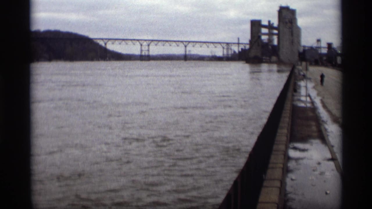 1969 Saint Paul riverfront view with a bridge, industrial silo structure, wet walkway, railing, snow patches, and one distant person.