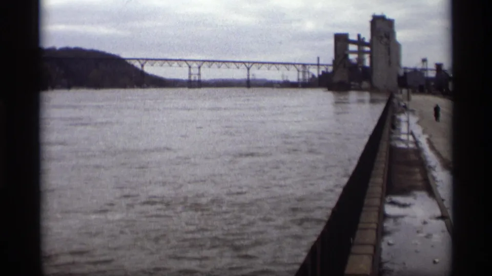 1969 home-movie frame of the Saint Paul riverfront, with a long bridge, industrial silos, railing, wet pavement, snow, and distant figure.