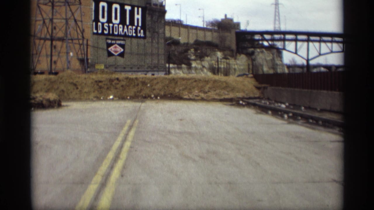 1969 Saint Paul 8mm view of a quiet road approaching an industrial storage sign, rail track, embankment, and bridge.