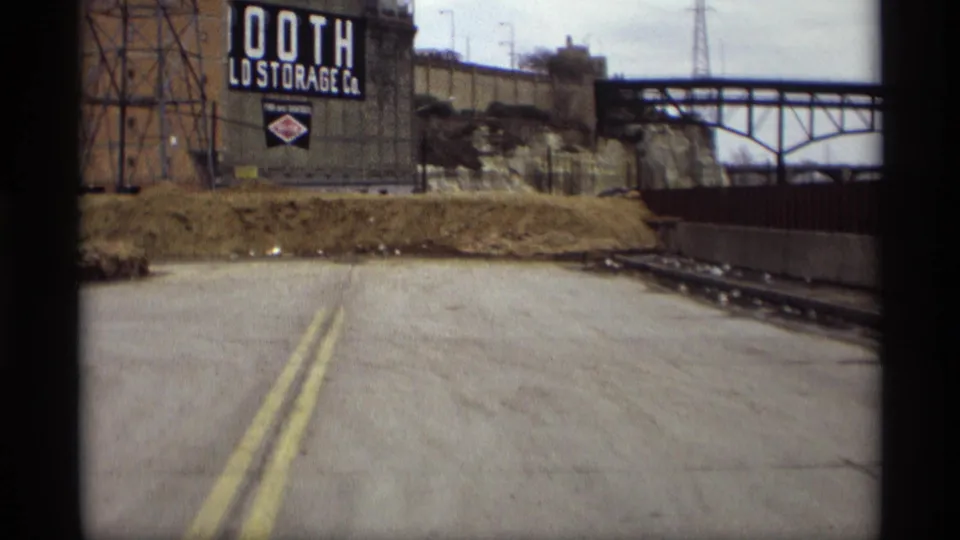 1969 Saint Paul 8mm frame showing a road curve area, industrial storage sign, rail corridor, and bridge over the river area.