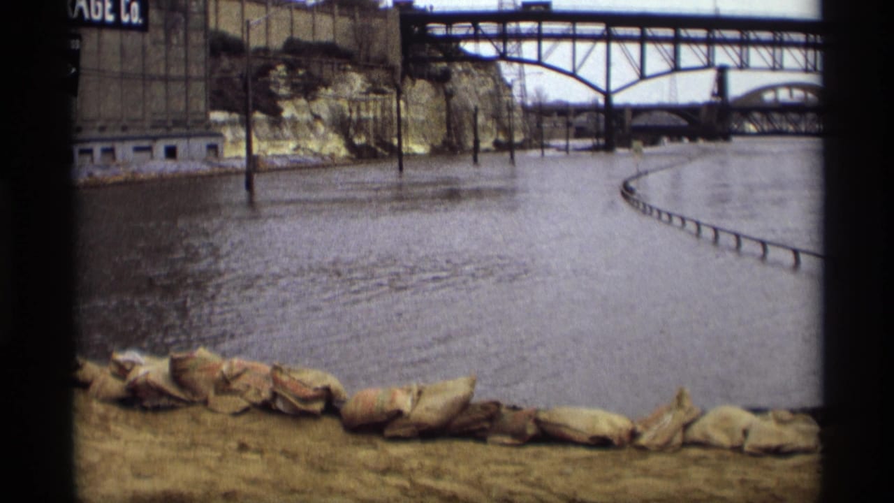 Floodwater beside sandbags with industrial buildings and a steel bridge in the background.