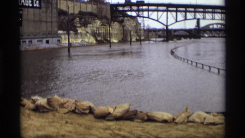 1969 Saint Paul floodwater presses near sandbags, with a steel truss bridge and industrial buildings visible beyond.