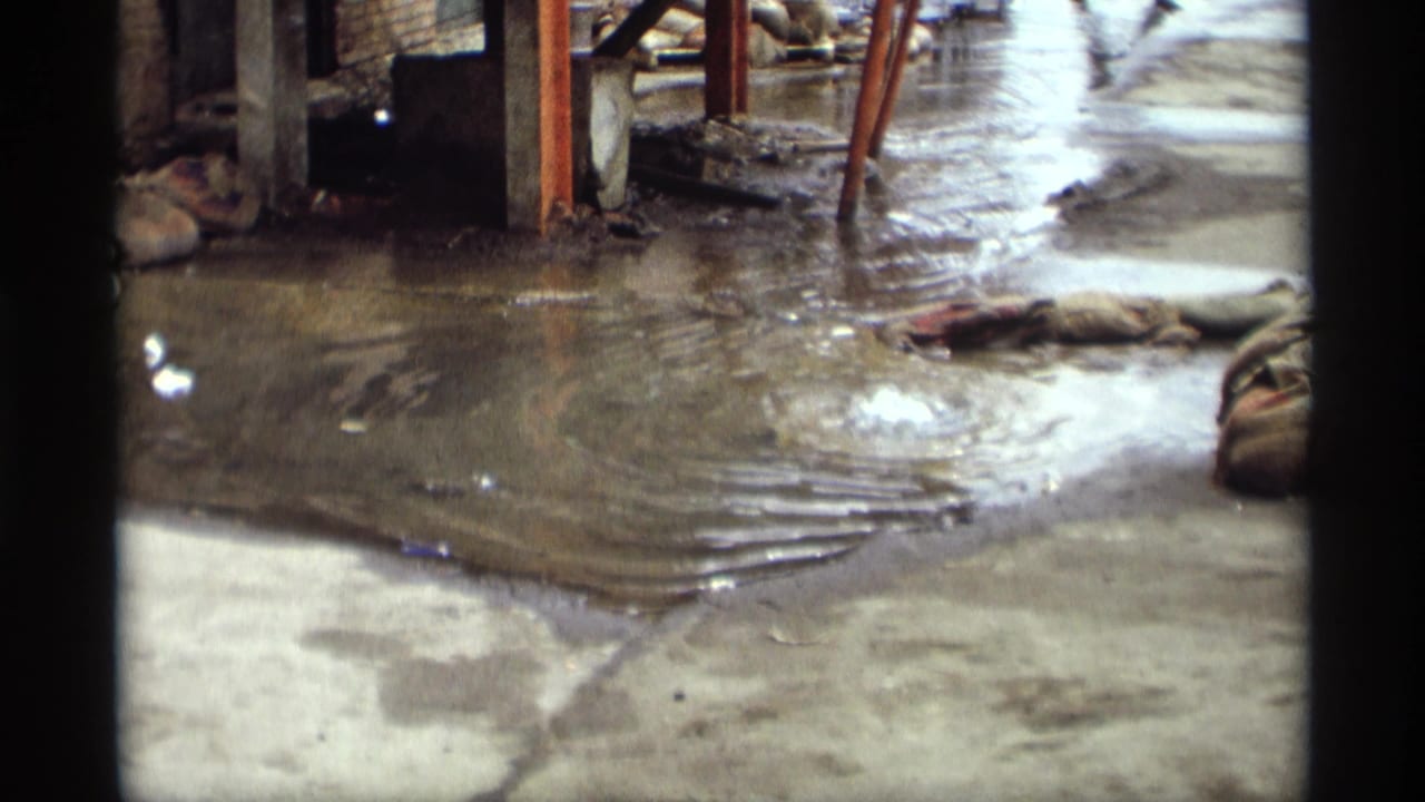 1969 Saint Paul 8mm frame showing rippling murky water on concrete, with dark frame edges, metal supports, and scattered debris.
