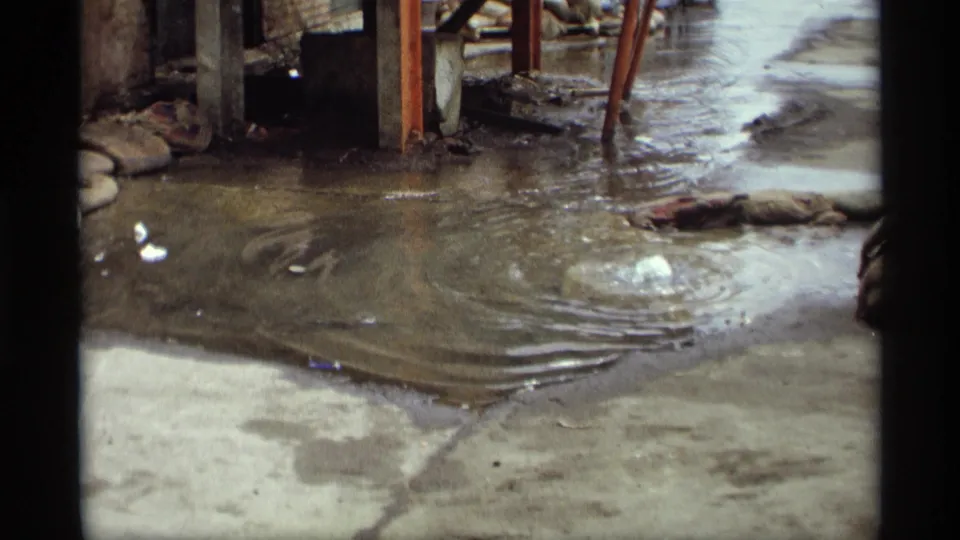 1969 Saint Paul frame of murky rippling water on a concrete surface, with a small bubble, metal supports, dark borders, and debris.