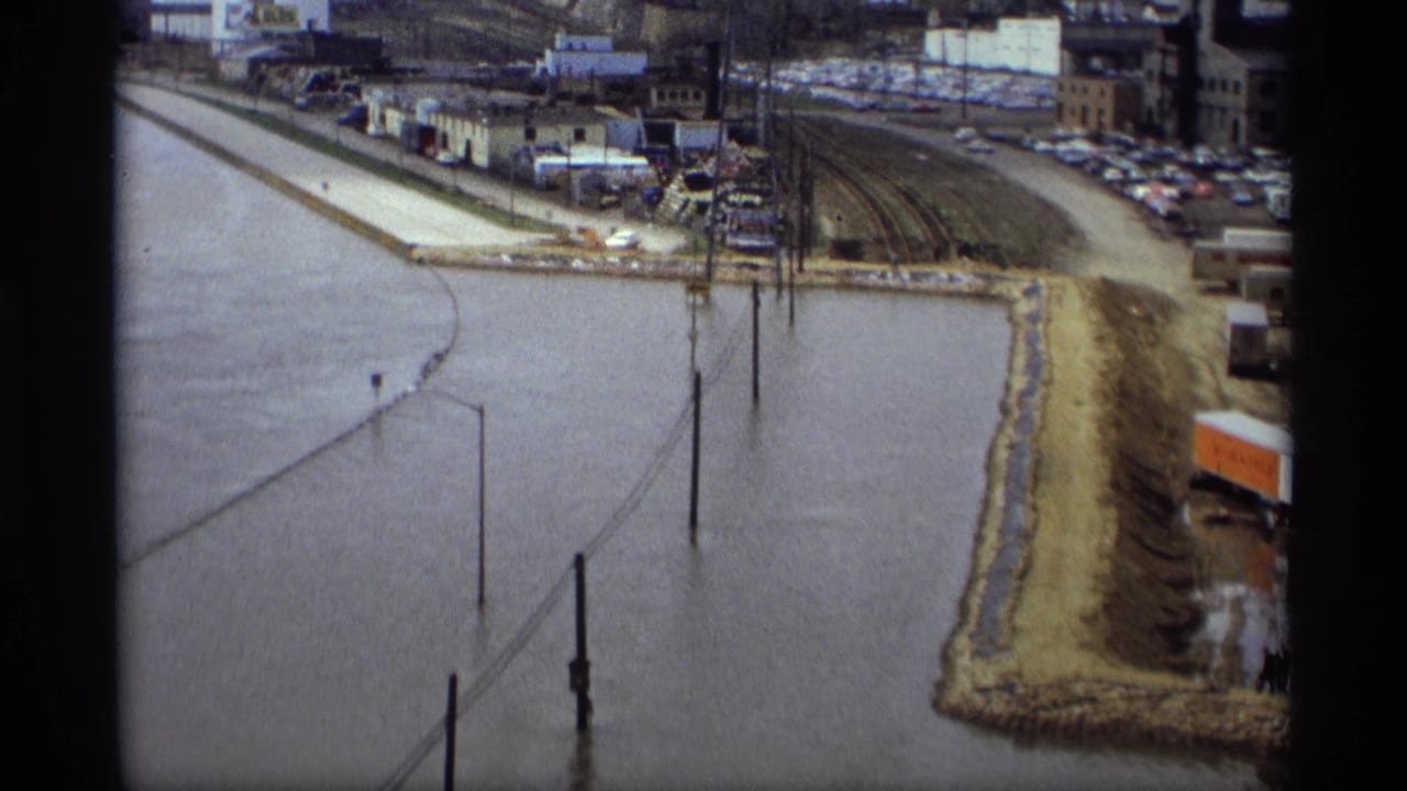 Floodwater covers a roadway with streetlights and utility poles near a levee and industrial area.