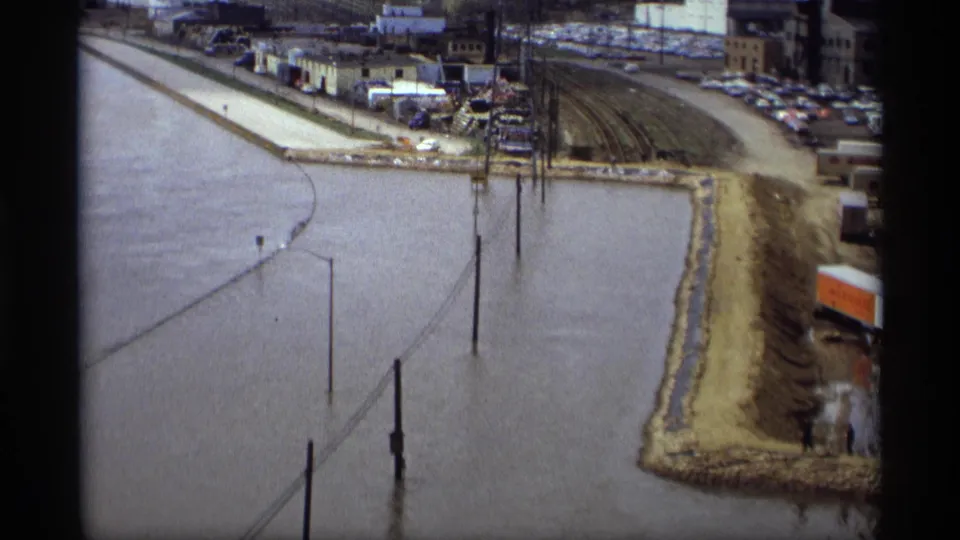 1969 Saint Paul flood footage showing muddy water against a levee, with submerged poles, rail tracks, parked cars, and industrial buildings beyond.